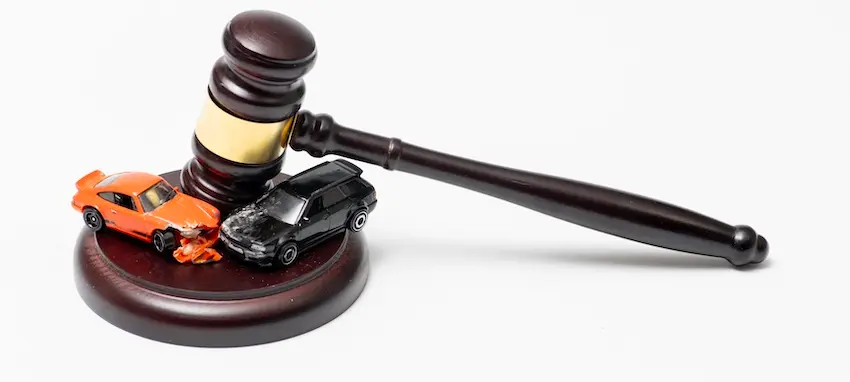 Two damaged toy cars- one orange and one black- are pinned beneath a wooden judge's gavel on its sound block against a white background symbolizing the legal action and court proceedings- with help from NC Injury Law- that follow rideshare accidents involving injured passengers or third parties.