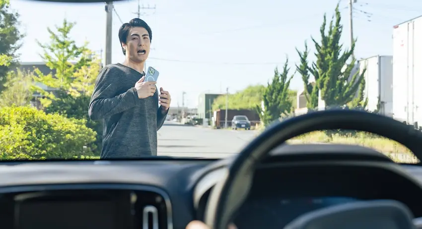 A shocked man holding a cell phone stands directly in front of an approaching vehicle viewed from inside the car's dashboard depicting a dangerous near-miss pedestrian encounter caused by a driver who failed to follow right-of-way laws in North Carolina.