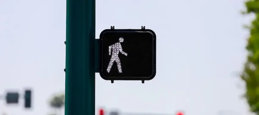 A pedestrian walk signal illuminated on a green pole at an intersection against a light blue/gray sky representing the traffic control signals that reinforce right-of-way laws designed to protect pedestrians crossing the street in North Carolina.