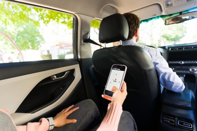 A passenger in the back seat of a car holds a smartphone displaying a rideshare app map while a driver in a button-down shirt sits in the front depicting the typical scenario in which rideshare accidents can occur during an active trip in North Carolina.