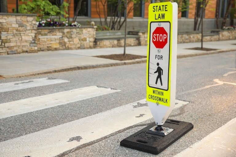 A bright yellow and white crosswalk sign in the center of a painted crosswalk reading State Law Stop for Pedestrians Within Crosswalk directly referencing the right-of-way laws that require drivers to yield to pedestrians in marked crossing zones in North Carolina.