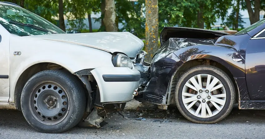 A white sedan and a dark navy vehicle with severely crumpled front ends are pressed against each other head-on near a large tree trunk on a paved street with broken plastic bumper debris scattered across the ground beneath both cars and a canopy of green trees visible in the soft-focus background — the kind of serious collision that typically gives rise to a car accident lawsuit with help from NC Injury Law.