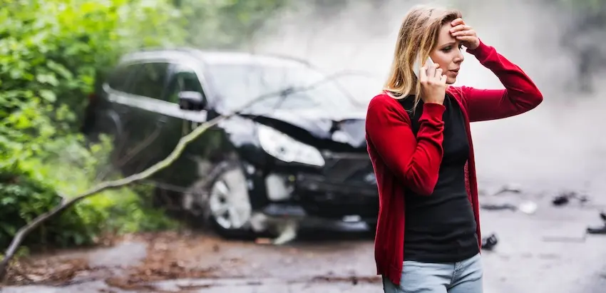 A visibly shaken blonde woman wearing a red zip-up jacket over a black shirt stands on a wet debris-covered road while talking on the phone with NC Injury Law and raising her other hand to her forehead in distress with a totaled black SUV behind her billowing white smoke from its crumpled hood amid overhanging green foliage — capturing the traumatic aftermath of a crash that would warrant pursuing a car accident lawsuit in North Carolina.