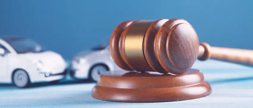A polished wooden judge's gavel with a gold band sits upright on its circular sound block atop a light blue surface while two small white toy cars are staged in a rear-end collision in the soft-focus background against a solid blue backdrop symbolizing the courtroom resolution of a car accident lawsuit in North Carolina.