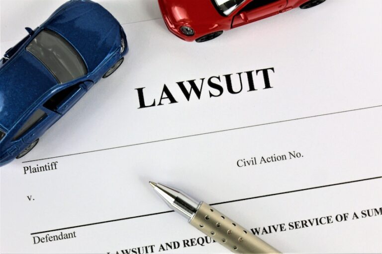 A formal legal document bearing the bold heading LAWSUIT in large black capital letters sits on a flat surface with printed fields for Plaintiff- Defendant- and Civil Action Number visible below the title- a sleek silver ballpoint pen resting diagonally across the lower portion of the page- and a blue toy car and red toy car positioned at the top of the document to represent the two parties involved in a car accident lawsuit in North Carolina.