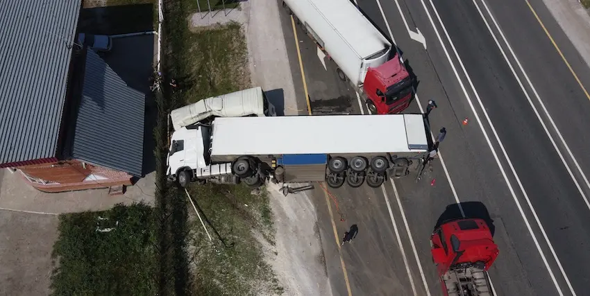 An aerial drone photograph looking straight down at a highway shoulder shows a large white semi-truck with a box trailer that has jackknifed and veered off the roadway- its cab angled sharply away from the trailer and coming to rest partially on a gravel shoulder beside a grassy strip and the metal roof of a gray industrial building while a red commercial truck is stopped on the right lane of the multi-lane highway with orange traffic cones visible on the road surface and two small figures of people standing near the scene — a wide-angle overhead view of the chaotic aftermath of a serious commercial vehicle accident in North Carolina.
