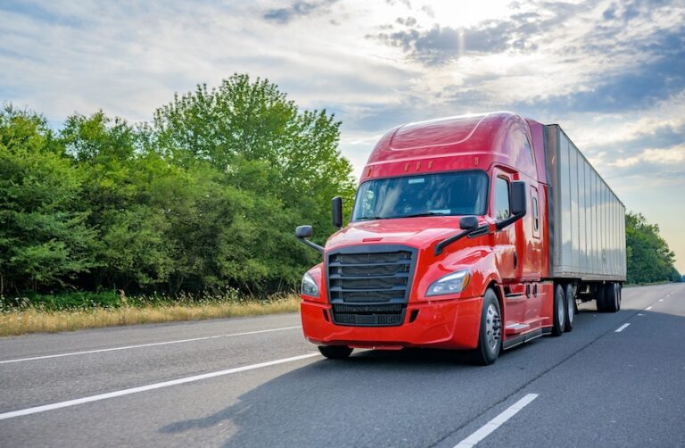 A massive red semi-truck with a sleek modern aerodynamic cab design and large black side mirrors pulls a long silver refrigerated trailer down a two-lane highway photographed head-on from a low angle that emphasizes its imposing size and forward momentum with a lush green tree line flanking both sides of the road and a dramatic partly cloudy sky with warm golden sunlight breaking through above — representing the type of large commercial trucking company vehicle that is frequently involved in a commercial vehicle accident due to its size- weight- and extended hours on the road in North Carolina.