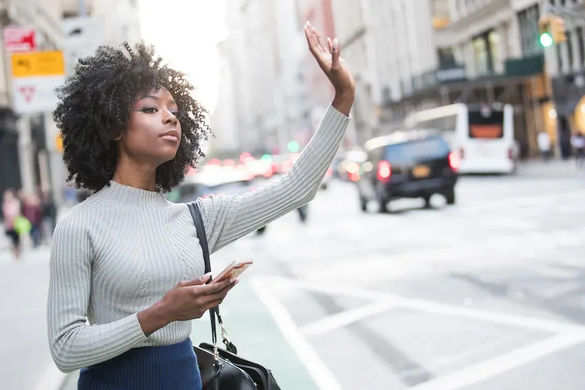 A woman raising her hand on the street to signal for a rideshare driver to pick her up.