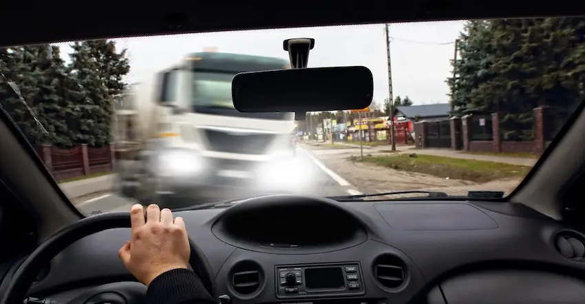 A first-person perspective shot from the driver's seat of a passenger car shows a single hand gripping the lower portion of the black steering wheel while through the windshield a large white commercial truck with its headlights blazing is captured in motion blur crossing directly into the car's lane on a narrow suburban road with evergreen trees- utility poles- roadside businesses- and residential fencing visible on both sides of the street under an overcast gray sky — a harrowing point-of-view image depicting the terrifying split second before a commercial vehicle accident occurs and could use help from NC Injury Law.
