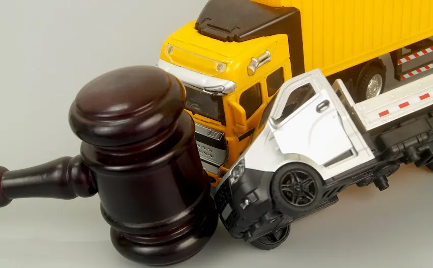 A dark mahogany judge's gavel- at NC Injury Law-with a wide rounded head stands upright in the left foreground against a neutral gray background positioned beside a staged collision scene featuring a bright yellow toy truck that has driven into the side of a white toy passenger car causing its door to swing open and the vehicle to be pushed sideways — a legal concept image using scale models to represent the liability and courtroom proceedings that follow a commercial vehicle accident involving a large truck and a smaller passenger vehicle in North Carolina.